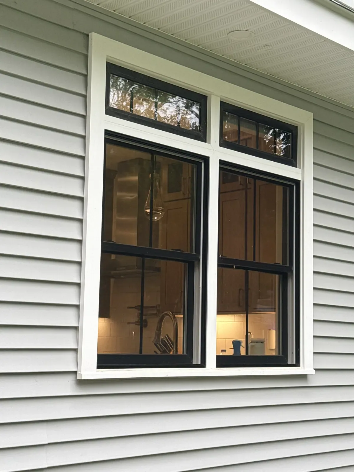 Twin double-hung windows with divided-lite transoms above, black frames and white trim, kitchen visible through the glass